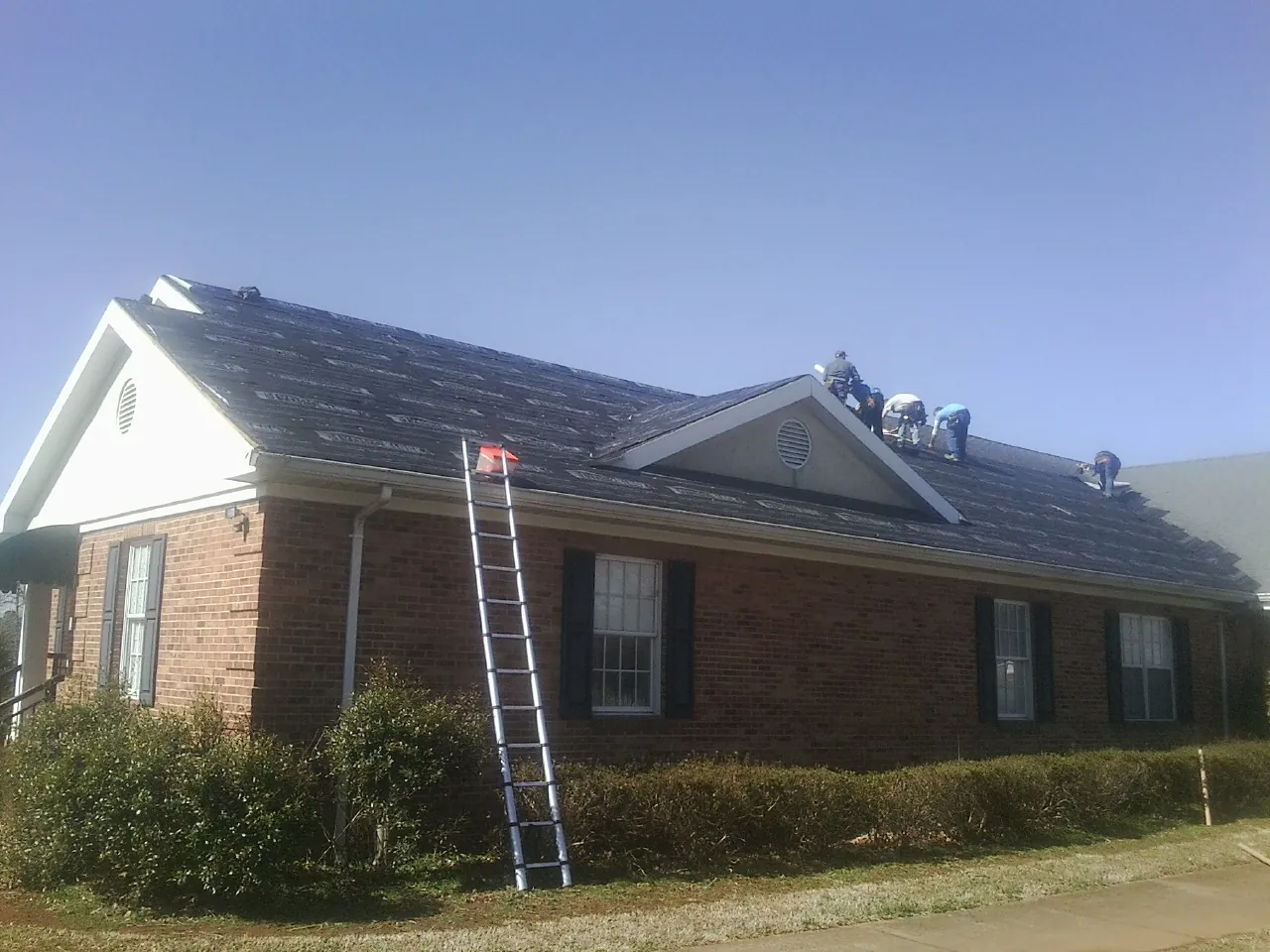 Crew tearing shingles off a brick ranch roof with a ladder set — Emergency Roof Repair work by our crew in Cranbury
