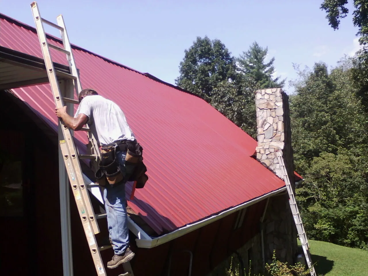 Roofer on a ladder beside a red metal roof and stone chimney — Soffit & Fascia Repair by Roofing Brothers in Cranbury